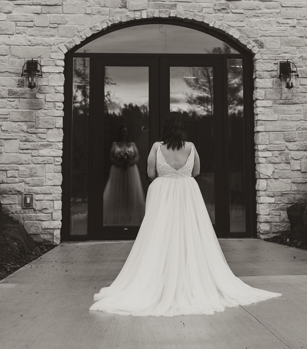 Black and white photo of a bride in a flowing gown standing before arched stone doorway reflecting in glass doors.