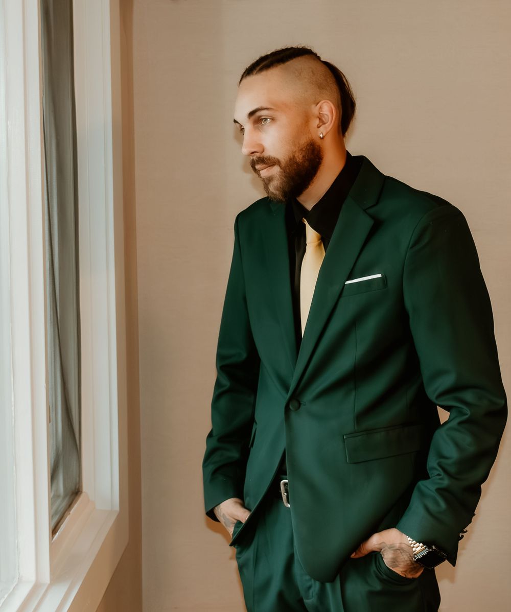 Stylish groom in dark green suit with gold tie gazes thoughtfully out a bright window.