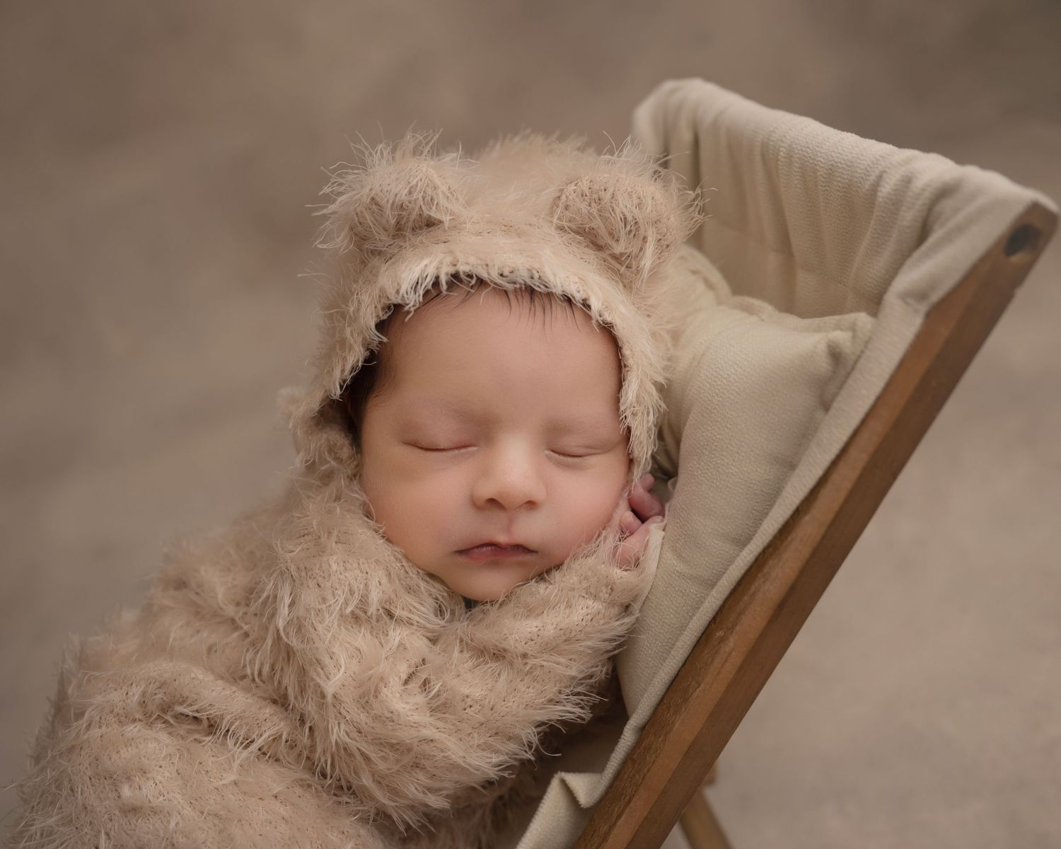 A sleeping baby in a fuzzy bear outfit rests in a beige wooden prop chair.