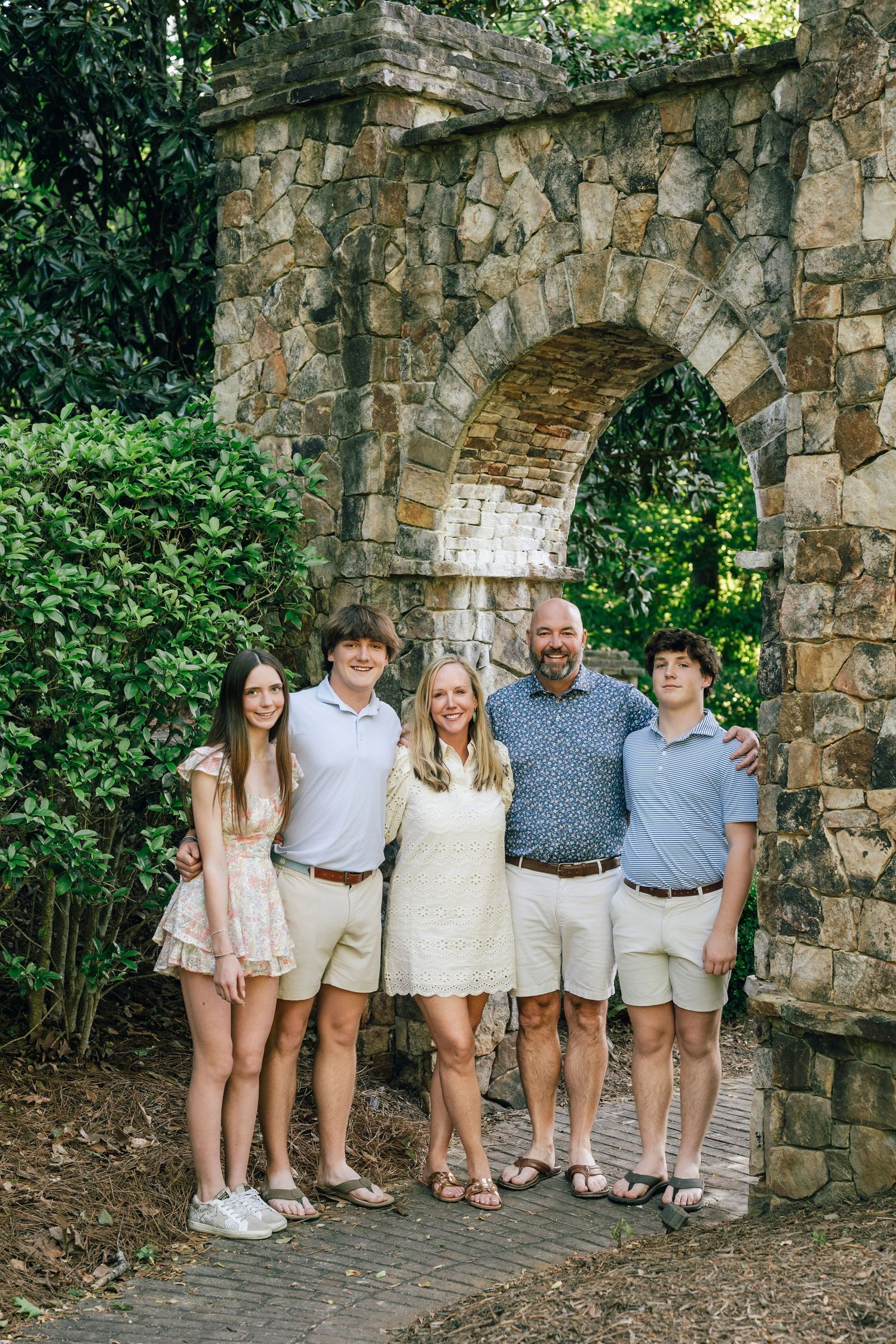 Family of five posing in front of a stone arch surrounded by lush greenery in a garden setting.