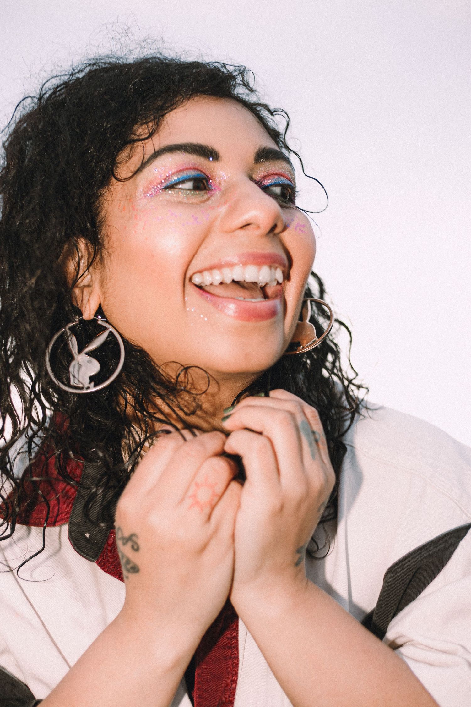 Sunshine with her joyful expression, dark curly hair, Playboy hoop earrings laughing against a white background.
