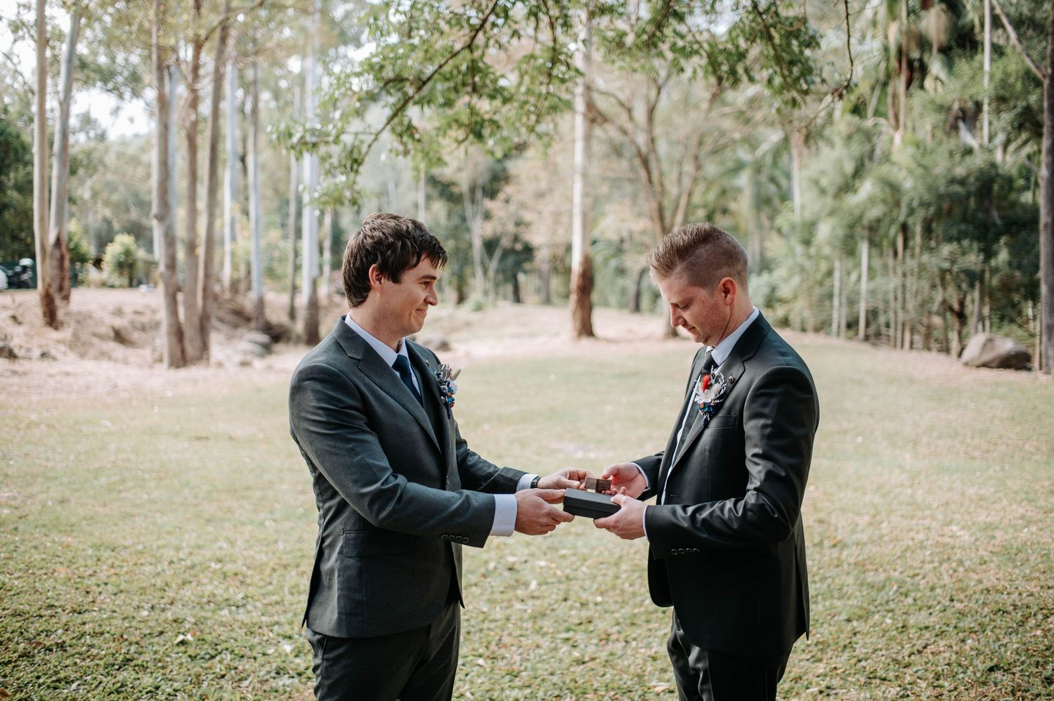 Two people in suits exchange rings during an outdoor wedding ceremony.
