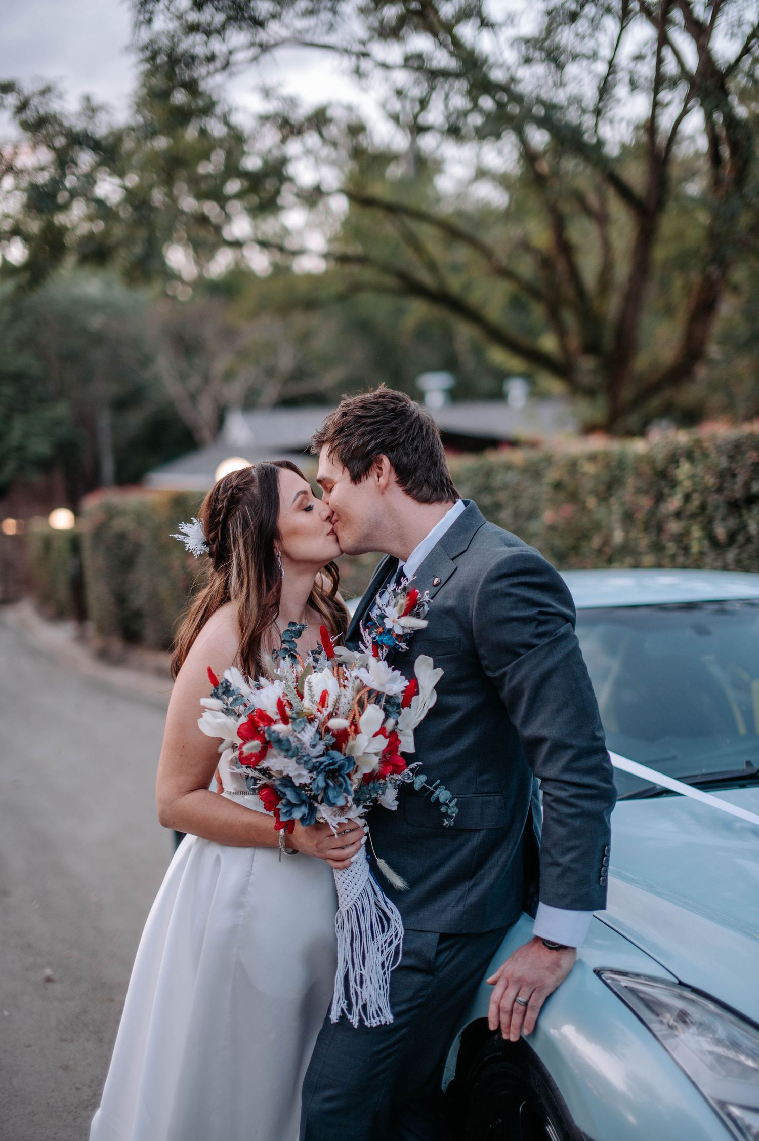 Tender moment between couple with red and blue bouquet beside parked car.