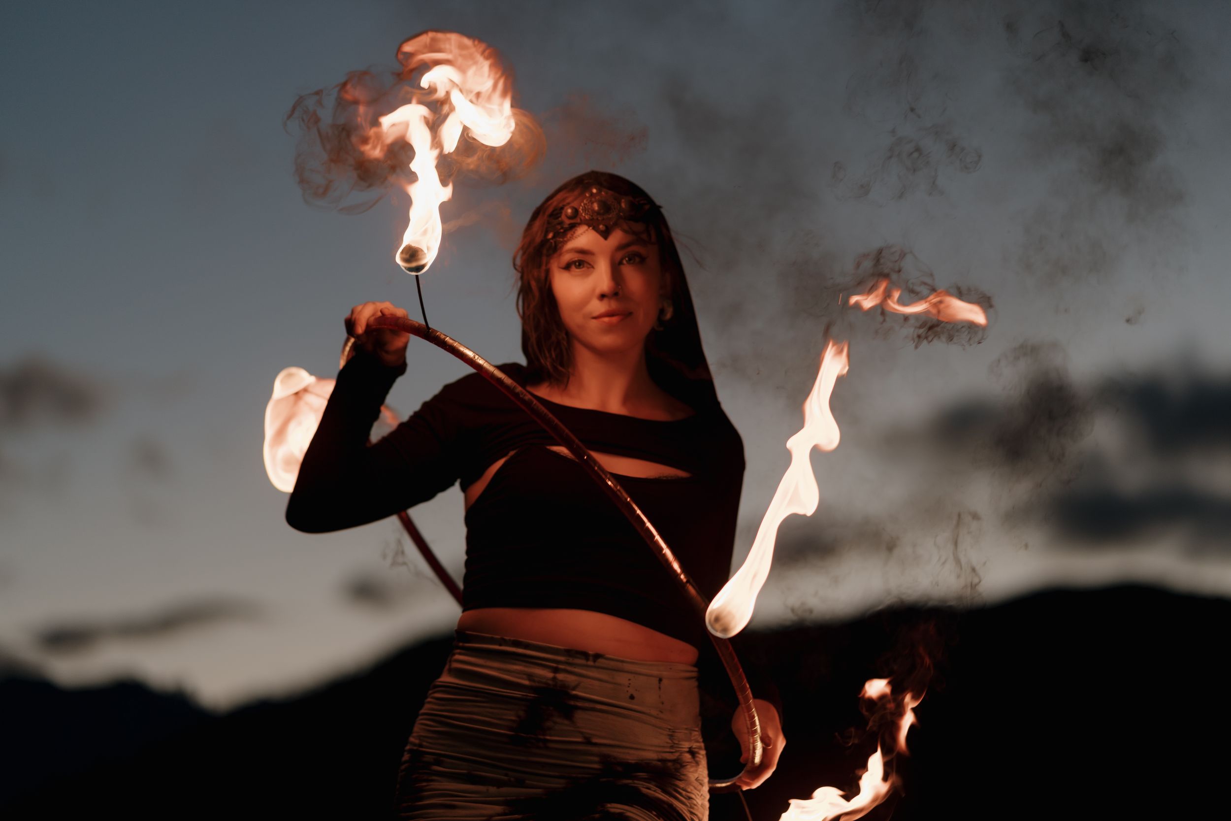Fire performer twirls flaming batons against a dark evening sky creating dramatic light trails.