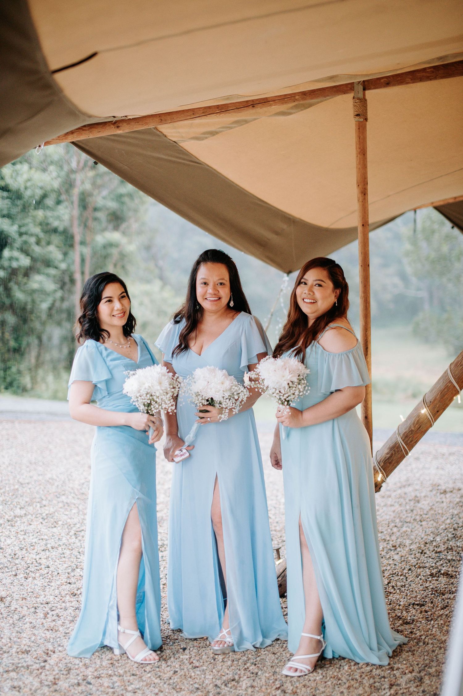 Three bridesmaids in light blue dresses holding bouquets under a tent.