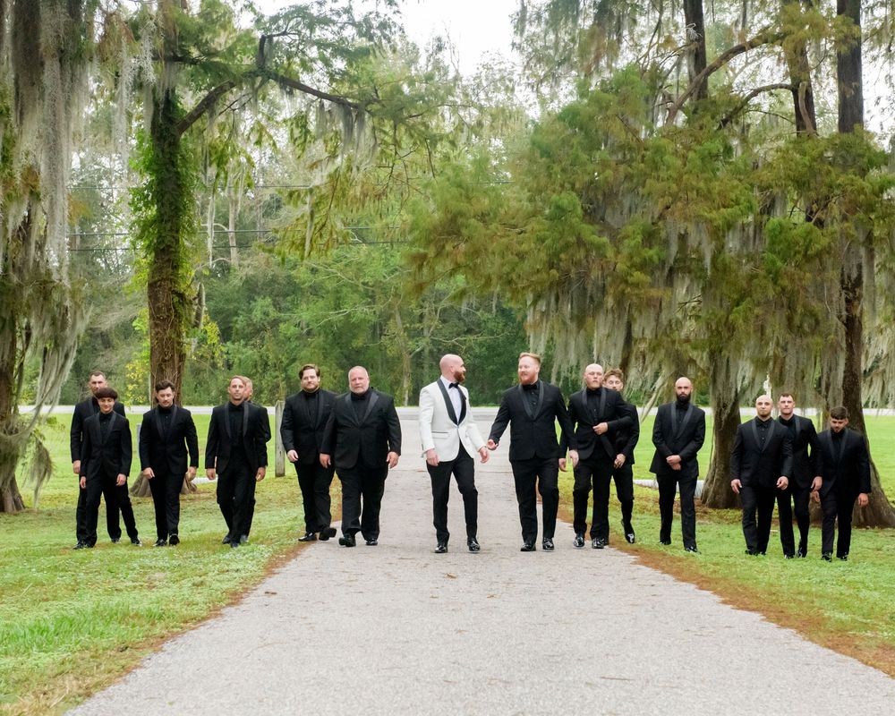 Wedding party wearing black and white formal attire stands on a tree-lined path with Spanish moss in the background.