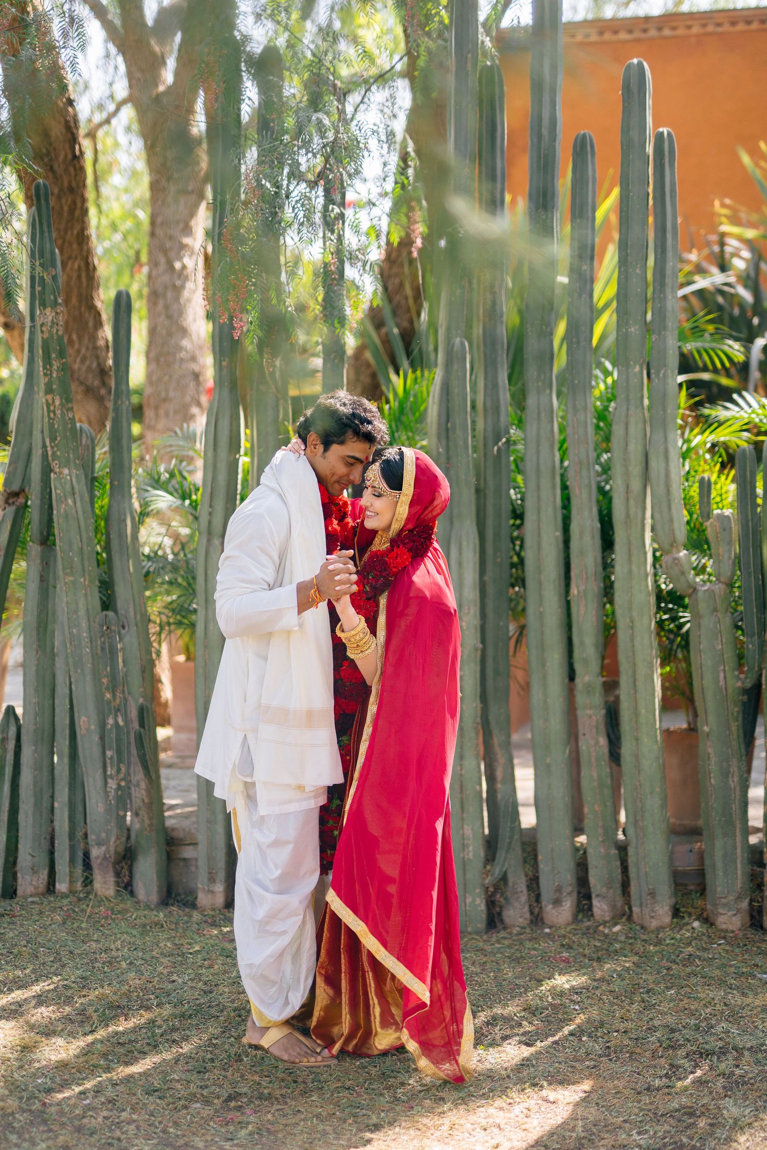 Couple in white and red traditional outfits sharing intimate moments among palm trees in a tropical garden.