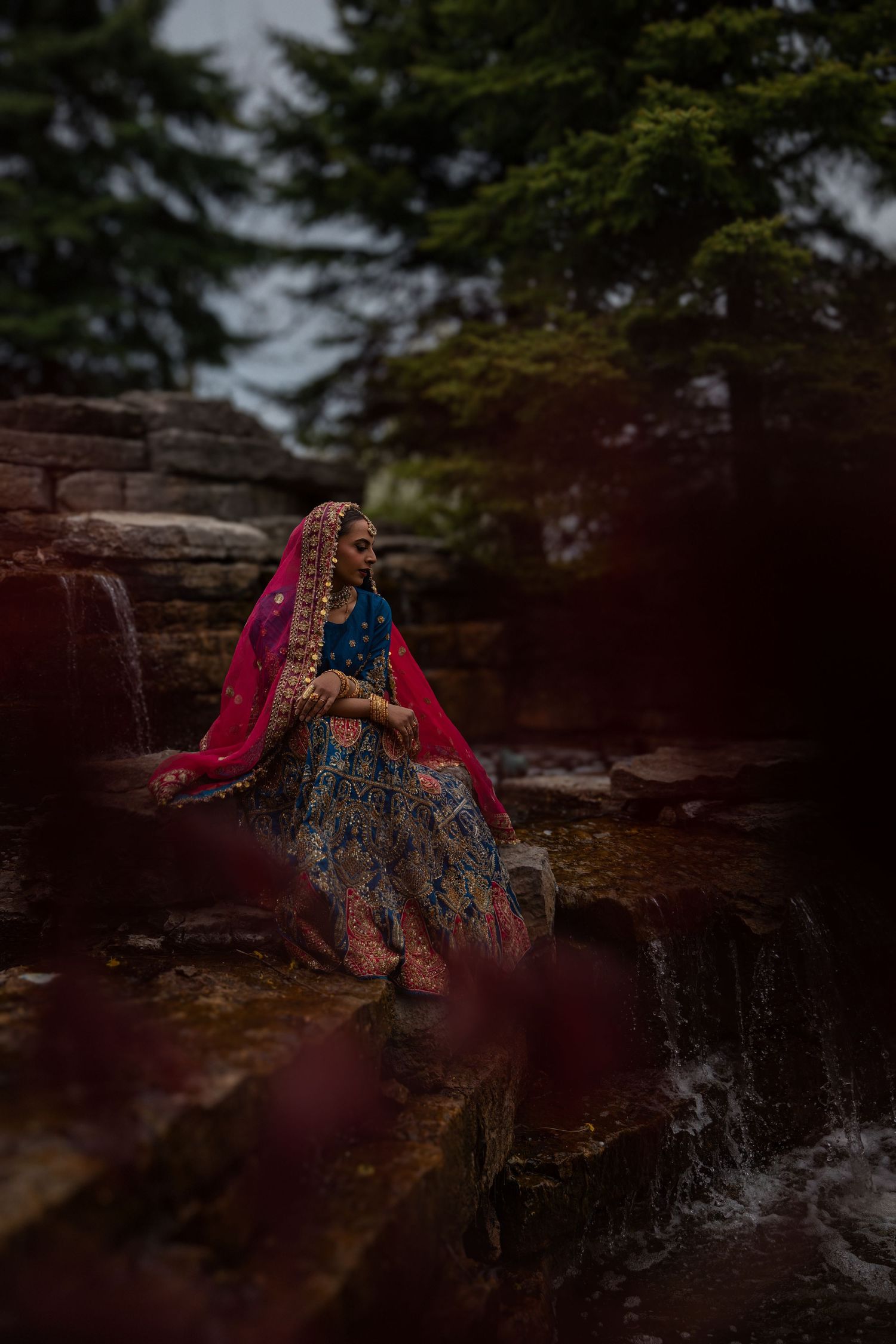 Elegant Black woman in blue shirt and paisley skirt sits on stone wall in colourful garden, smiling upward. Taken by BEXPRO in Brampton, ON.