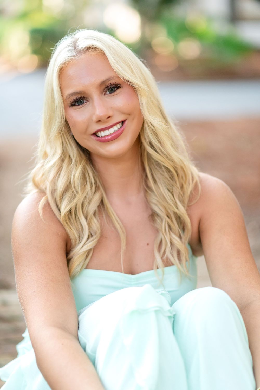 Smiling woman with long wavy blonde hair in a mint green dress sitting outdoors against a softly blurred background.