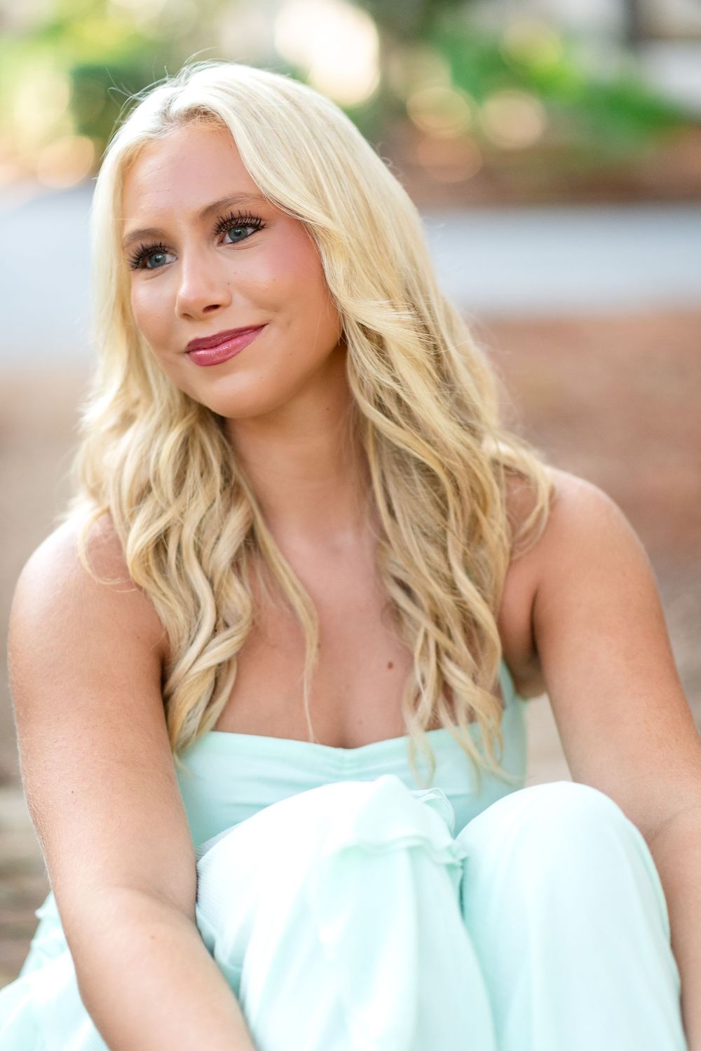 Smiling woman with long wavy blonde hair wearing a mint green strapless outfit, seated outdoors with a softly blurred background.