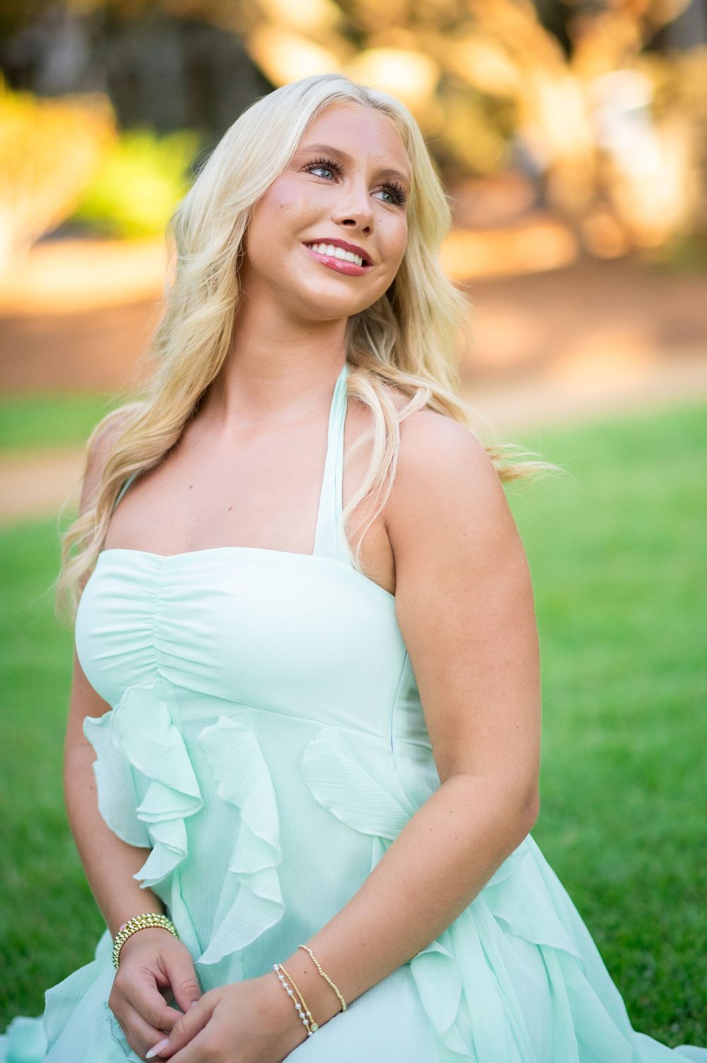 Smiling woman with long blonde hair in a mint green ruffled halter dress and gold bracelets sits on a sunlit grassy lawn outdoors.