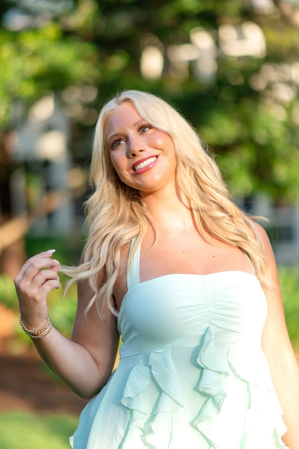 Smiling woman with long wavy blonde hair in a mint green ruffled sundress twirls a strand of hair outdoors among sunlit green trees.