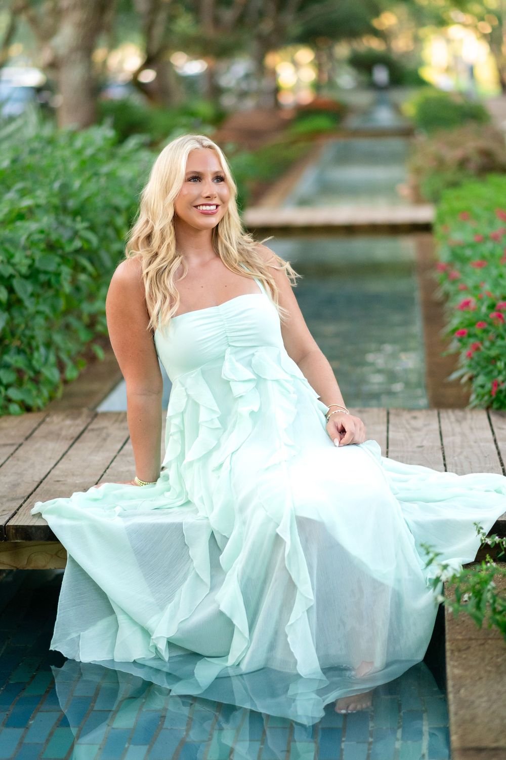 Blonde woman in a flowing mint green ruffled gown sits on a wooden boardwalk beside a reflecting pool surrounded by lush greenery.
