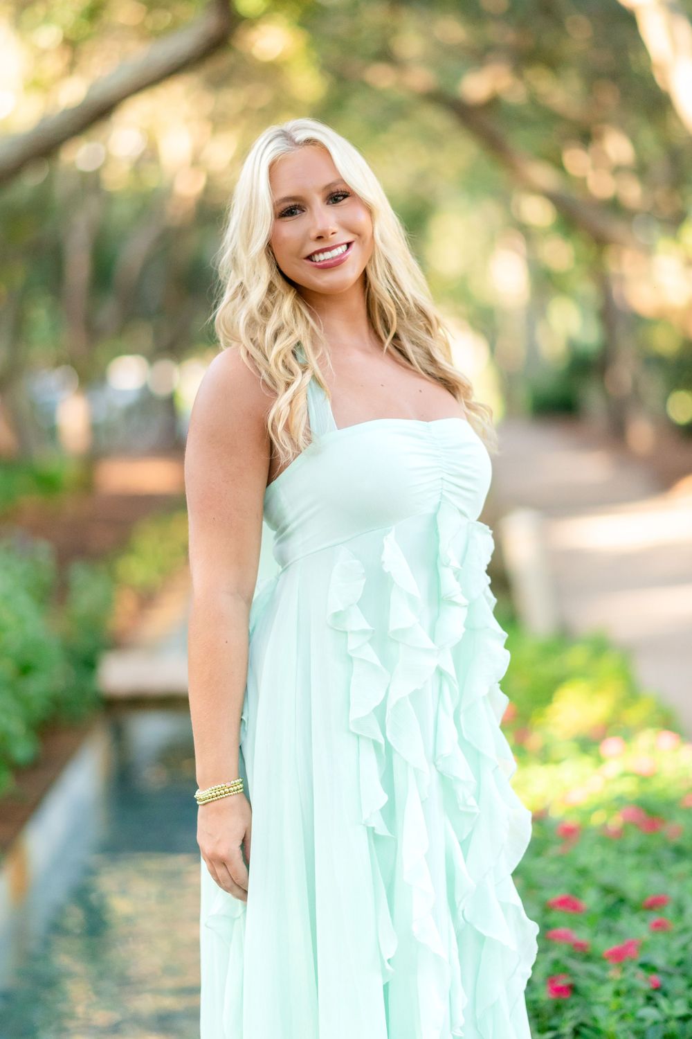 Smiling woman with long blonde wavy hair wearing a mint green ruffled halter dress standing in a lush, sunlit garden path.