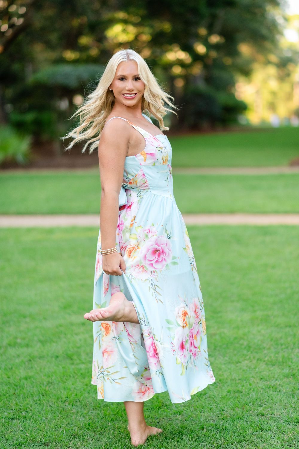 Smiling woman with long blonde hair in a light blue floral midi dress stands barefoot on green grass with one foot raised.