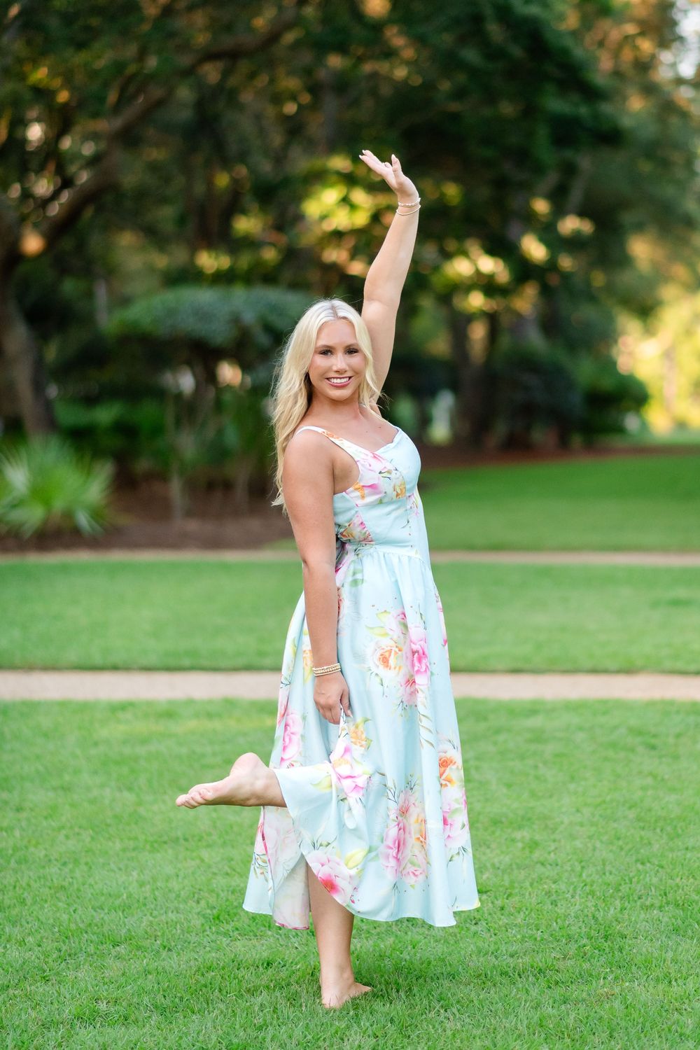 Smiling blonde woman in a floral pastel midi dress strikes a barefoot dance pose with one arm raised on a green lawn framed by trees.