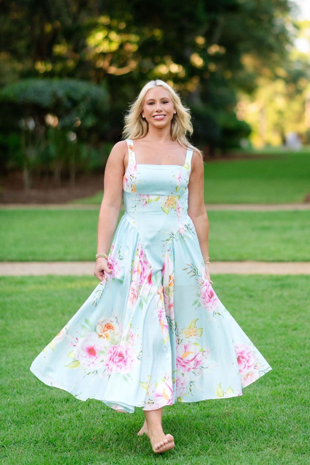 Woman with blonde hair twirls in a mint green floral midi sundress with square neckline and full skirt while walking on a sunny lawn.