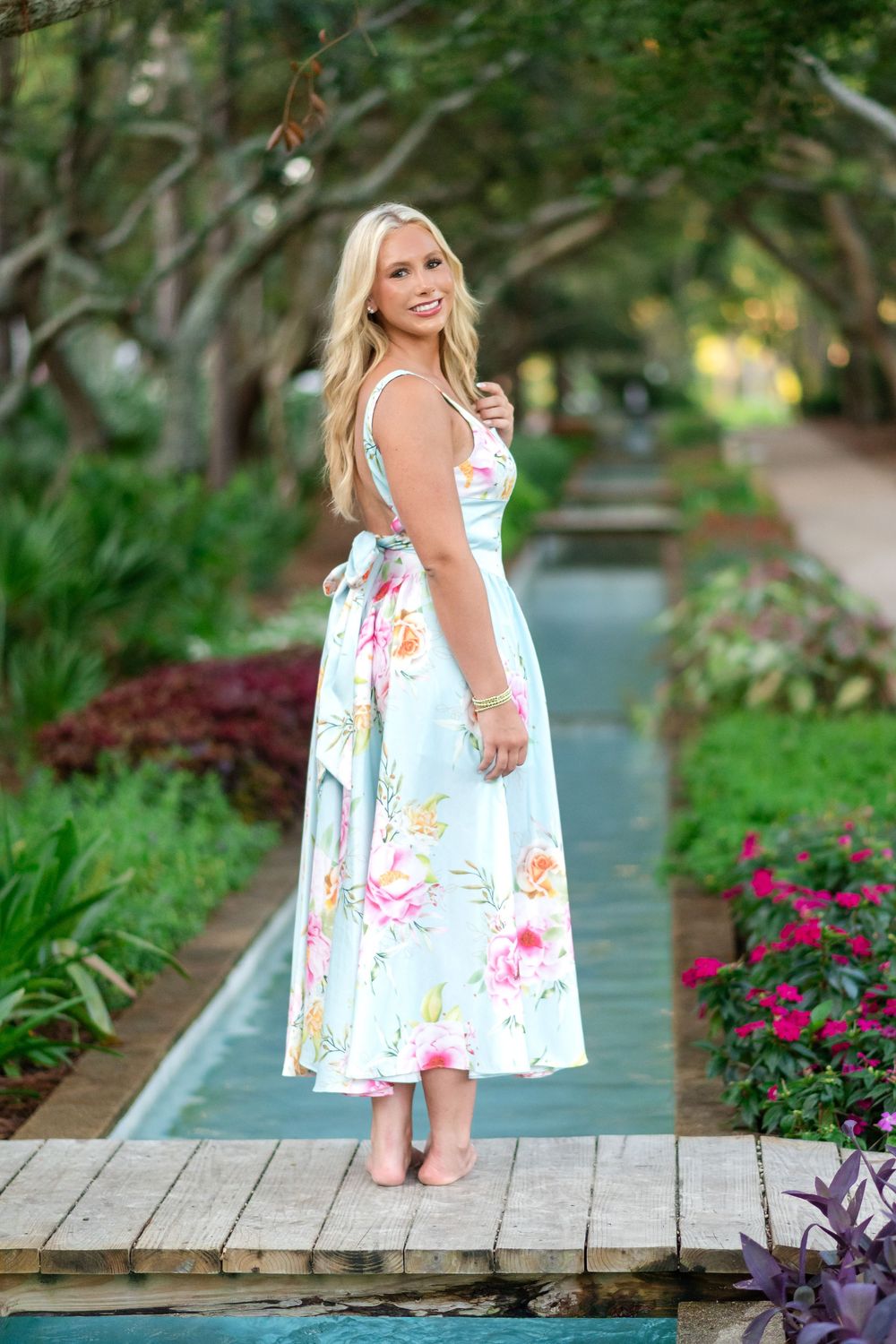 Blonde woman in a pastel floral midi dress standing on a wooden bridge over a garden water channel lined with pink flowers.