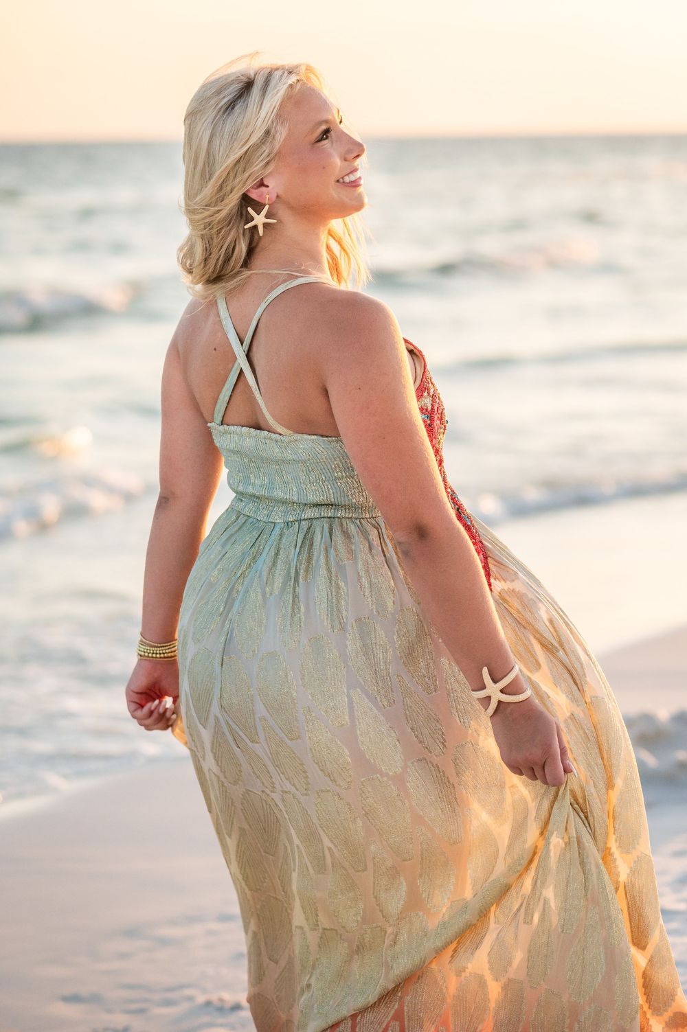 Smiling blonde woman in a flowing sage green dress and starfish earrings standing on the beach as the sun sets over the ocean.