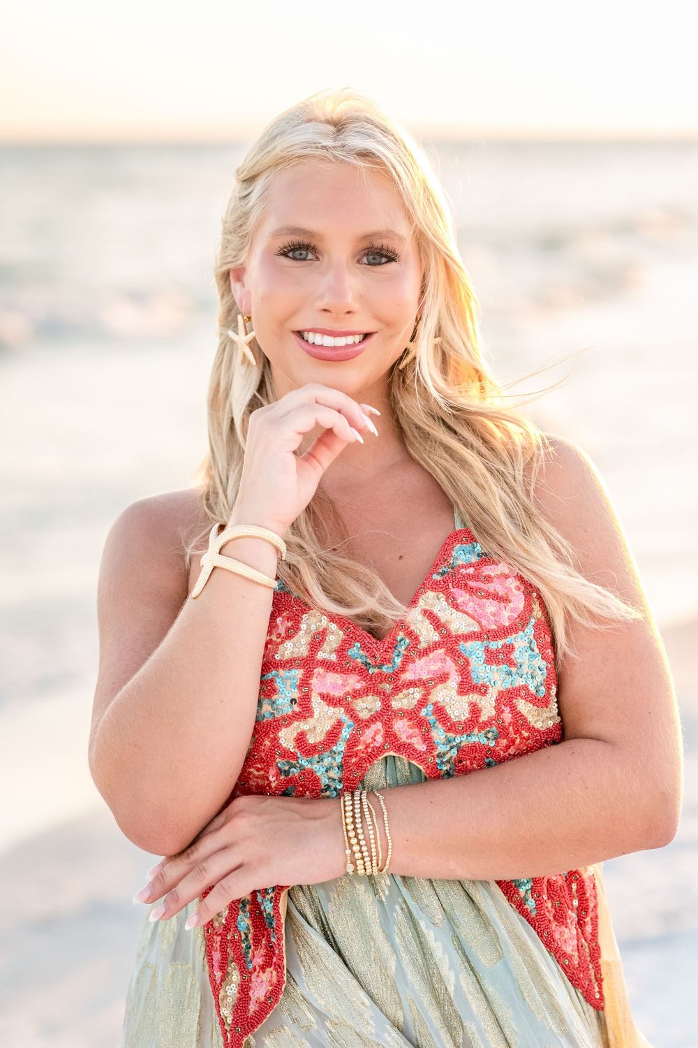 Smiling woman with long blonde hair poses on a sunlit beach in a red beaded top and flowing metallic dress.