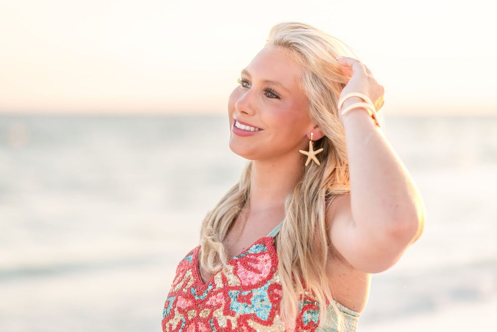 Smiling woman with long blonde hair and a starfish earring stands on a beach at sunset, wearing a colorful floral top.
