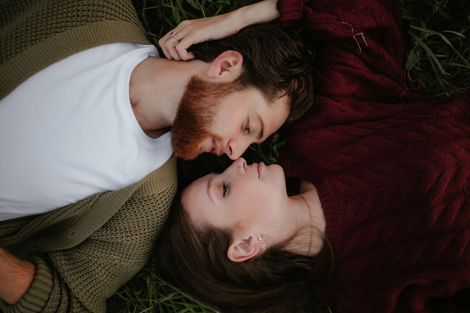 Overhead view of a bearded man and a woman lying nose to nose on the grass with eyes closed in a tender, romantic moment.