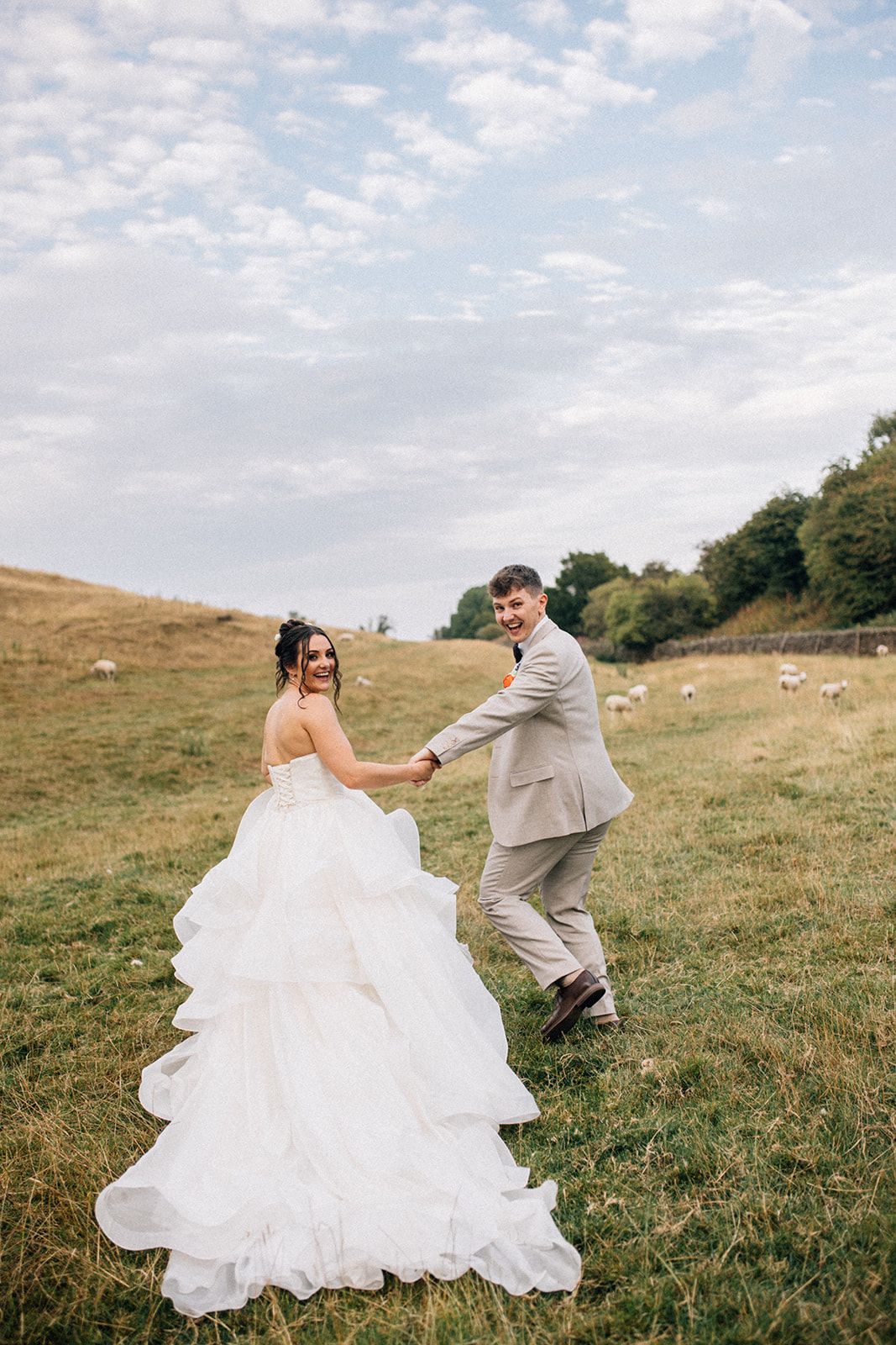 Bride in a ruffled white wedding gown and groom holding hands while walking through a grassy hillside field of the yorkshire dales