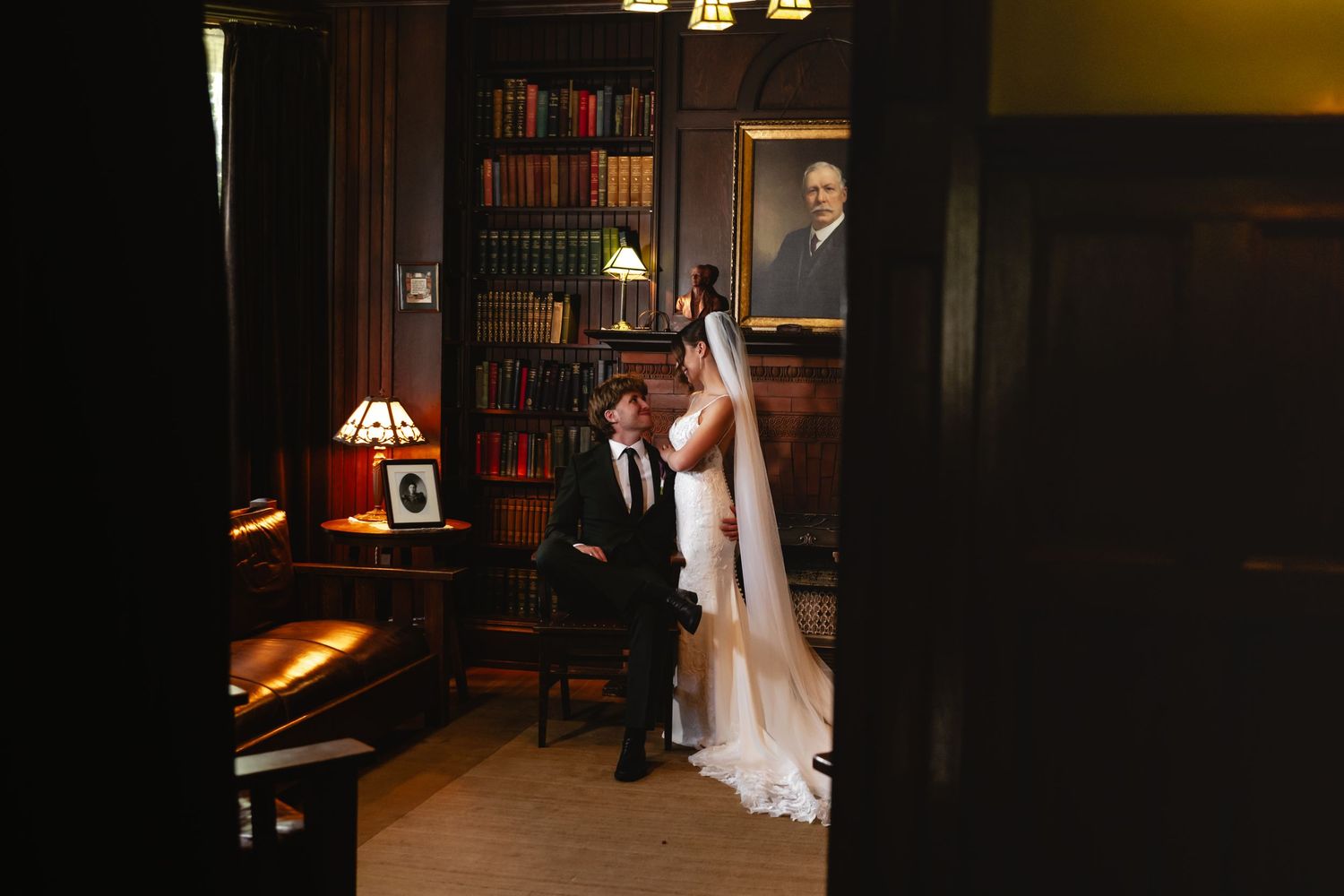 Bride in a white gown and long veil stands beside a seated groom in a dark suit in a cozy library with bookshelves and a framed portrait.