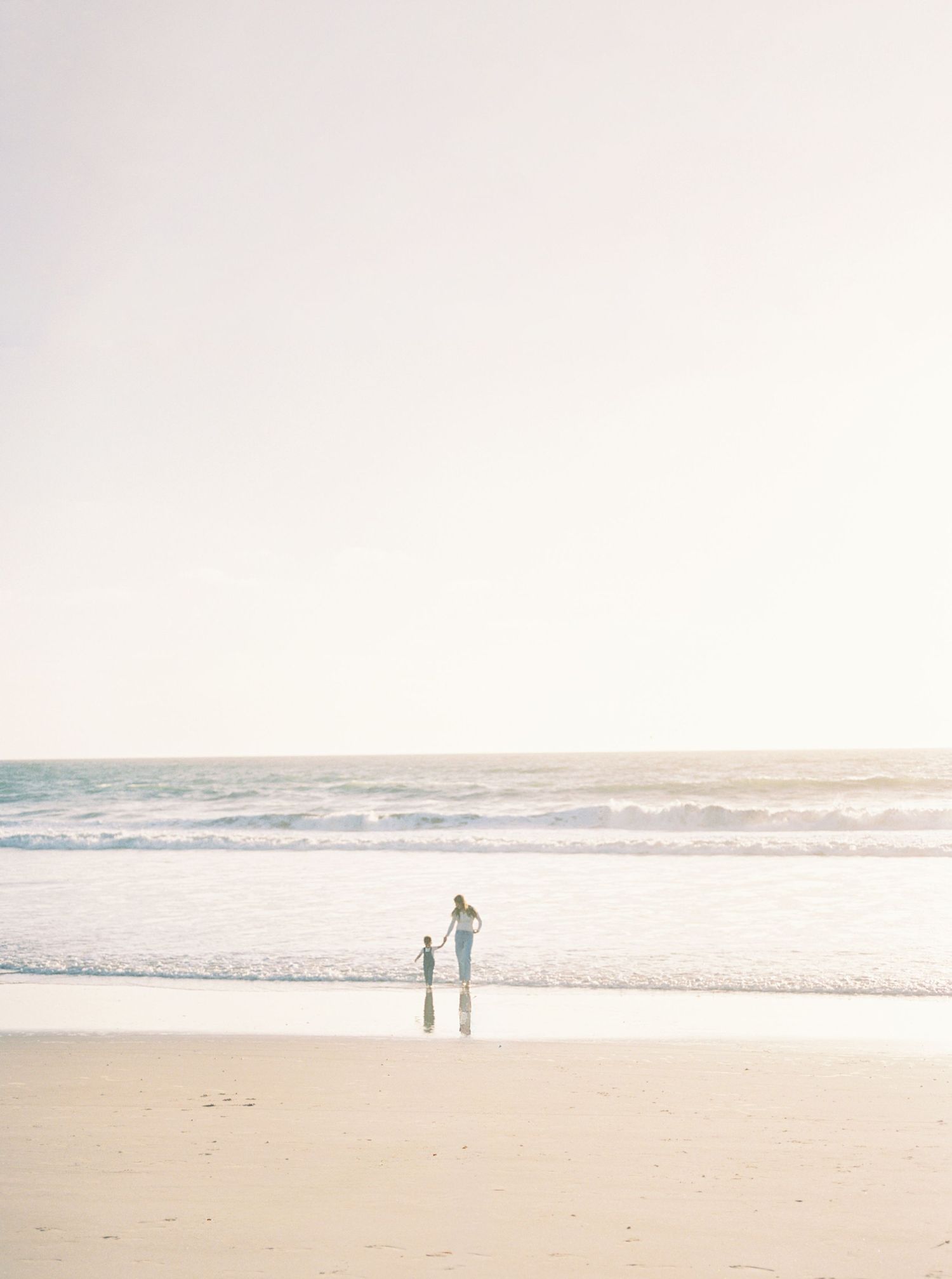Adult and child hold hands on a wide, sandy beach at sunset, with gentle waves rolling in under a soft, hazy sky.