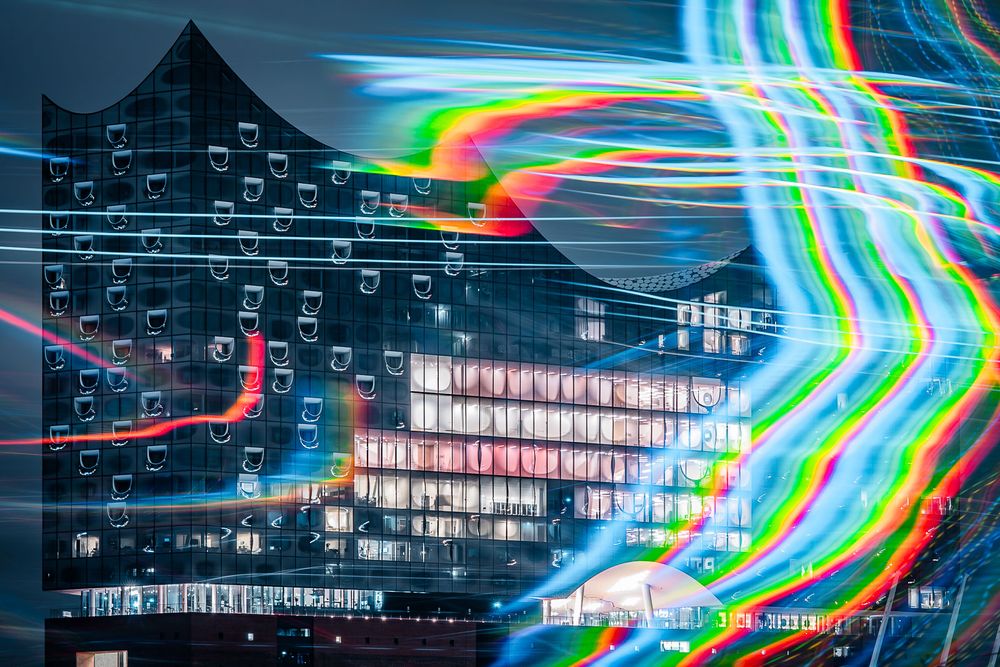 Long-exposure night photo of a modern glass concert hall with a wavy roofline, surrounded by vivid rainbow light trails.