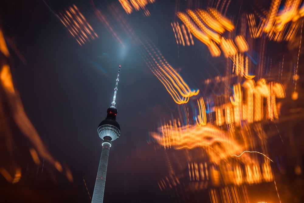 Berlin TV Tower rising into a dark night sky with streaking, blurred city lights creating colorful abstract light trails.