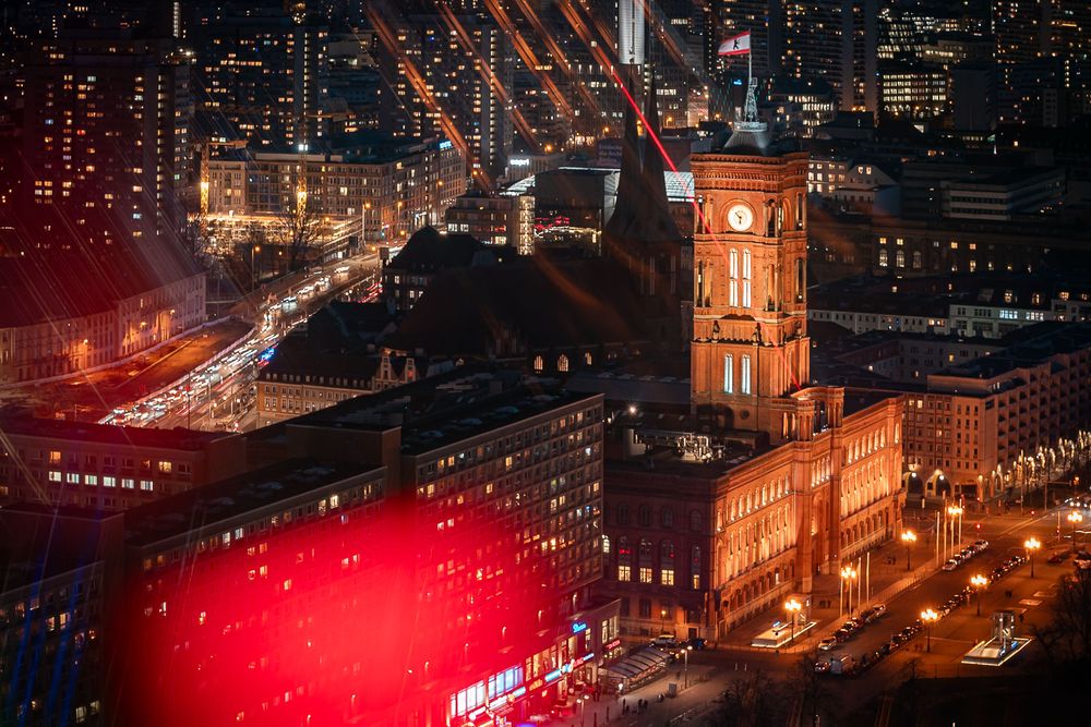 Aerial night view of a glowing red-brick city hall with a clock tower surrounded by illuminated buildings and streets.