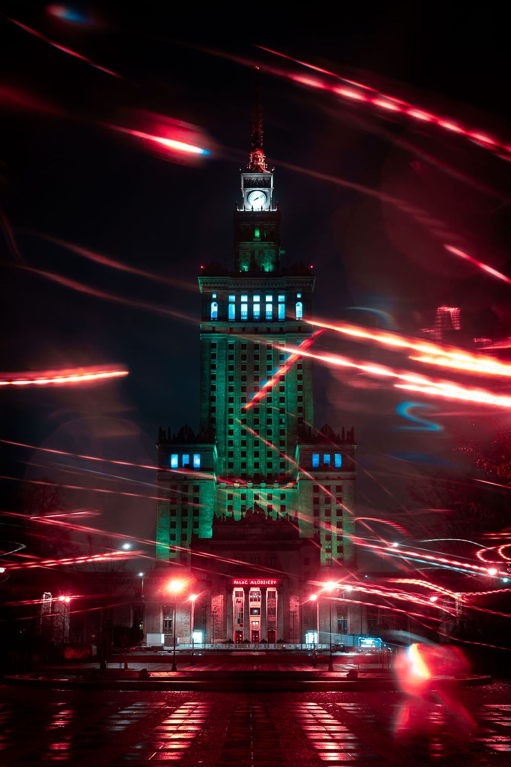 Illuminated Palace of Culture and Science skyscraper at night with red and blue light trails streaking across a dark Warsaw sky.
