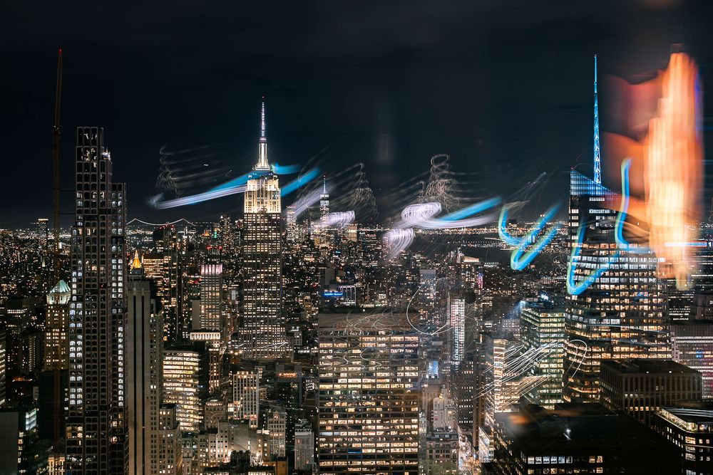 Long exposure night photo of the Manhattan skyline with the Empire State Building and glowing blue and orange light trails above the city.