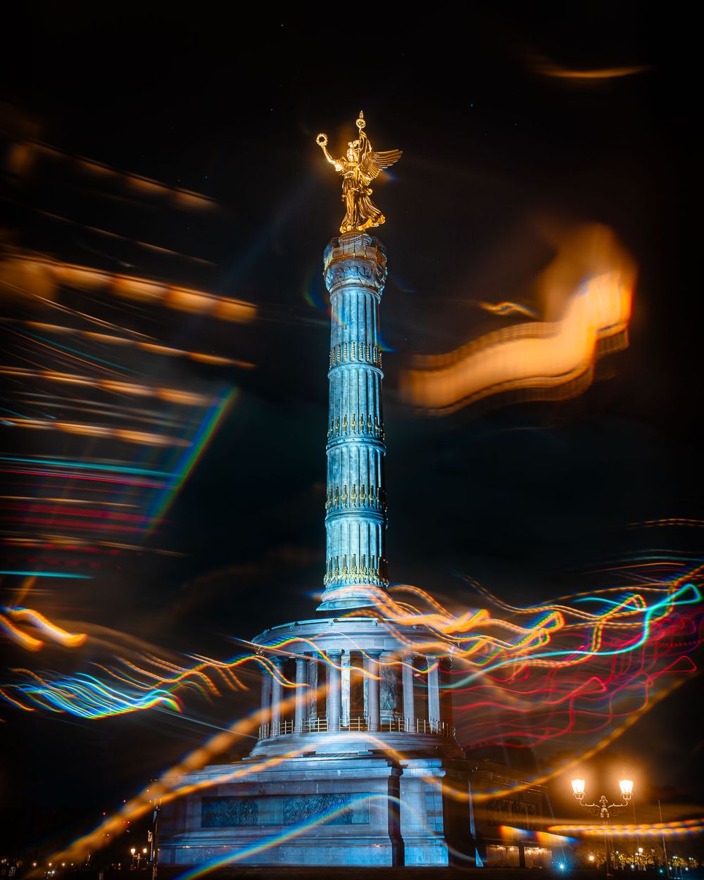 Illuminated Berlin Victory Column with golden statue at night, surrounded by colorful long-exposure light trails against a dark sky.