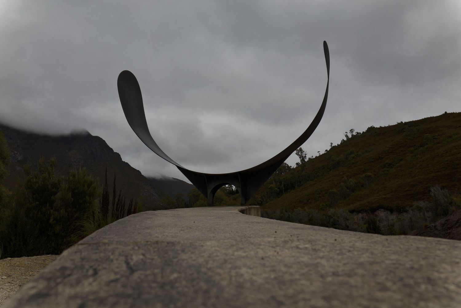 Black and white photo of a large curved metal sculpture with pointed tips standing on a rocky hillside under a cloudy sky.