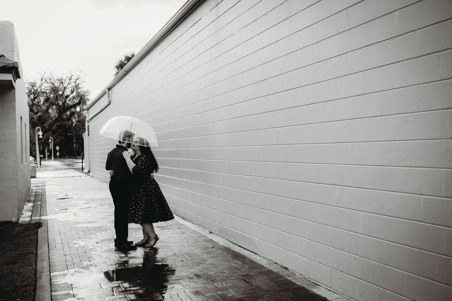 A couple embraces under a white umbrella on a rain-soaked sidewalk beside a tall wall in a moody black and white photo.