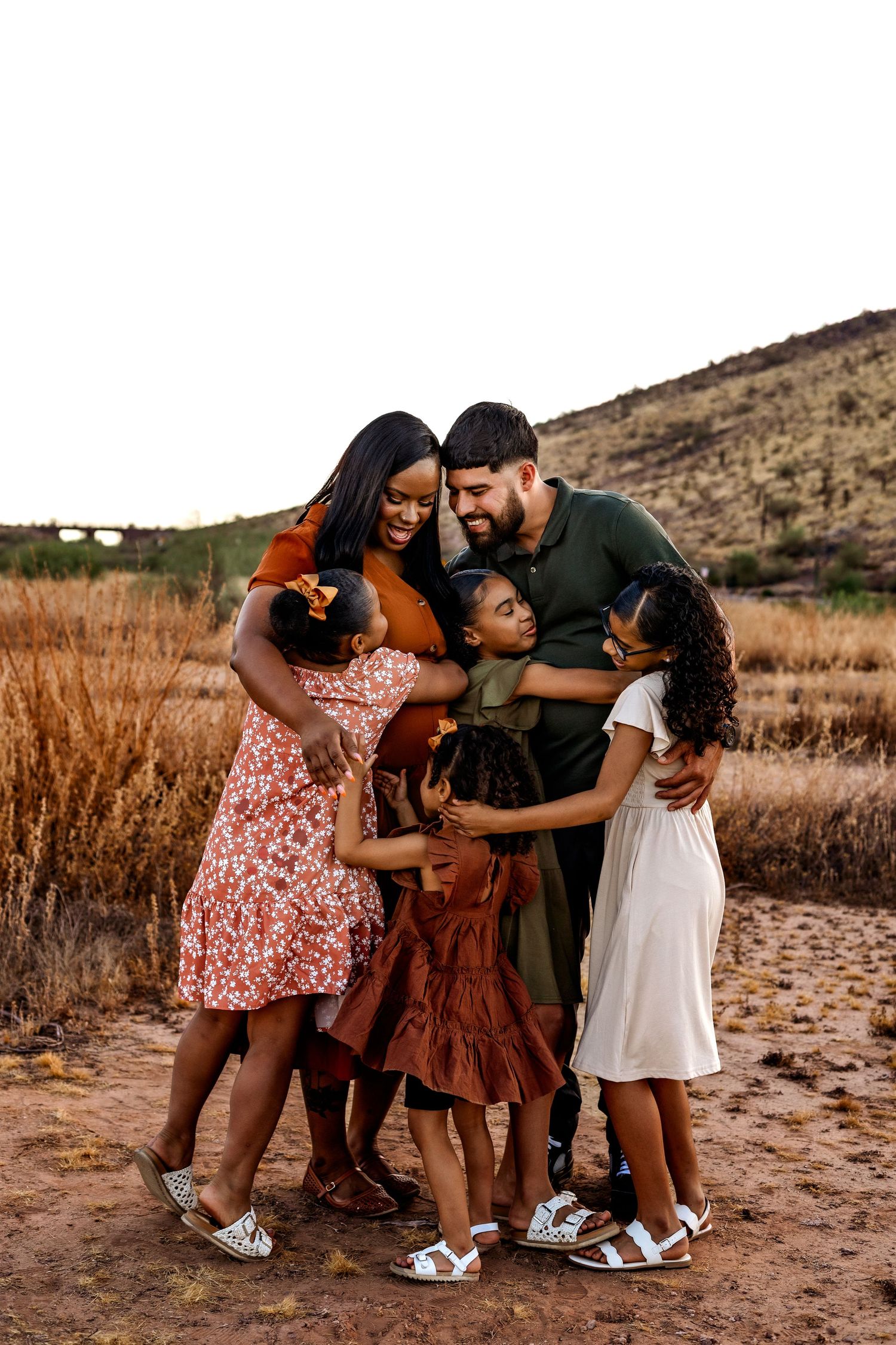 A joyful multiracial family of six shares a warm group hug outdoors in a golden desert landscape at sunset.