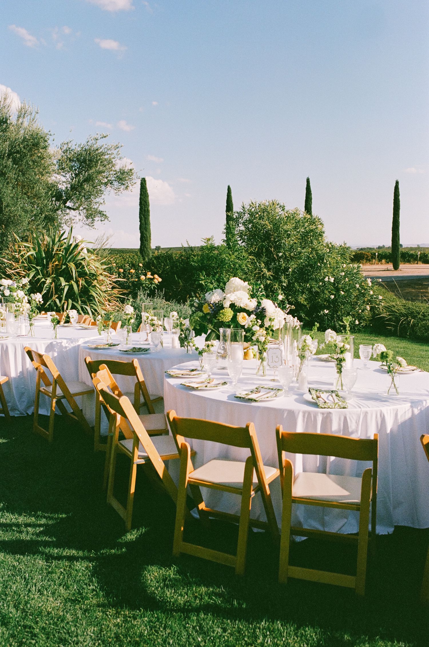 Elegant outdoor wedding reception with wooden folding chairs, white linen tables, floral centerpieces, and cypress trees.