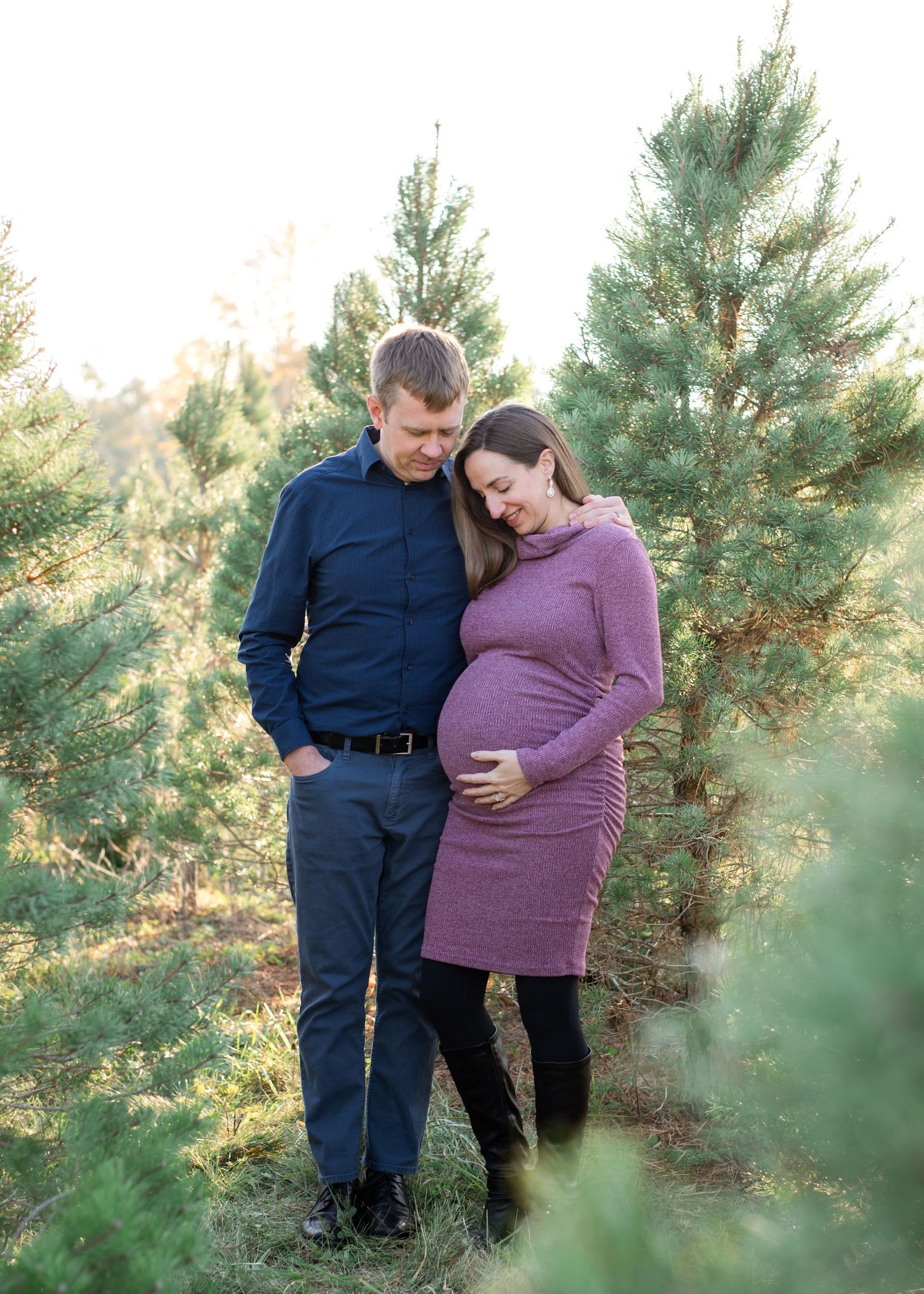 Smiling pregnant couple posing outdoors at a Christmas tree farm, woman in pink dress, man in plaid shirt.