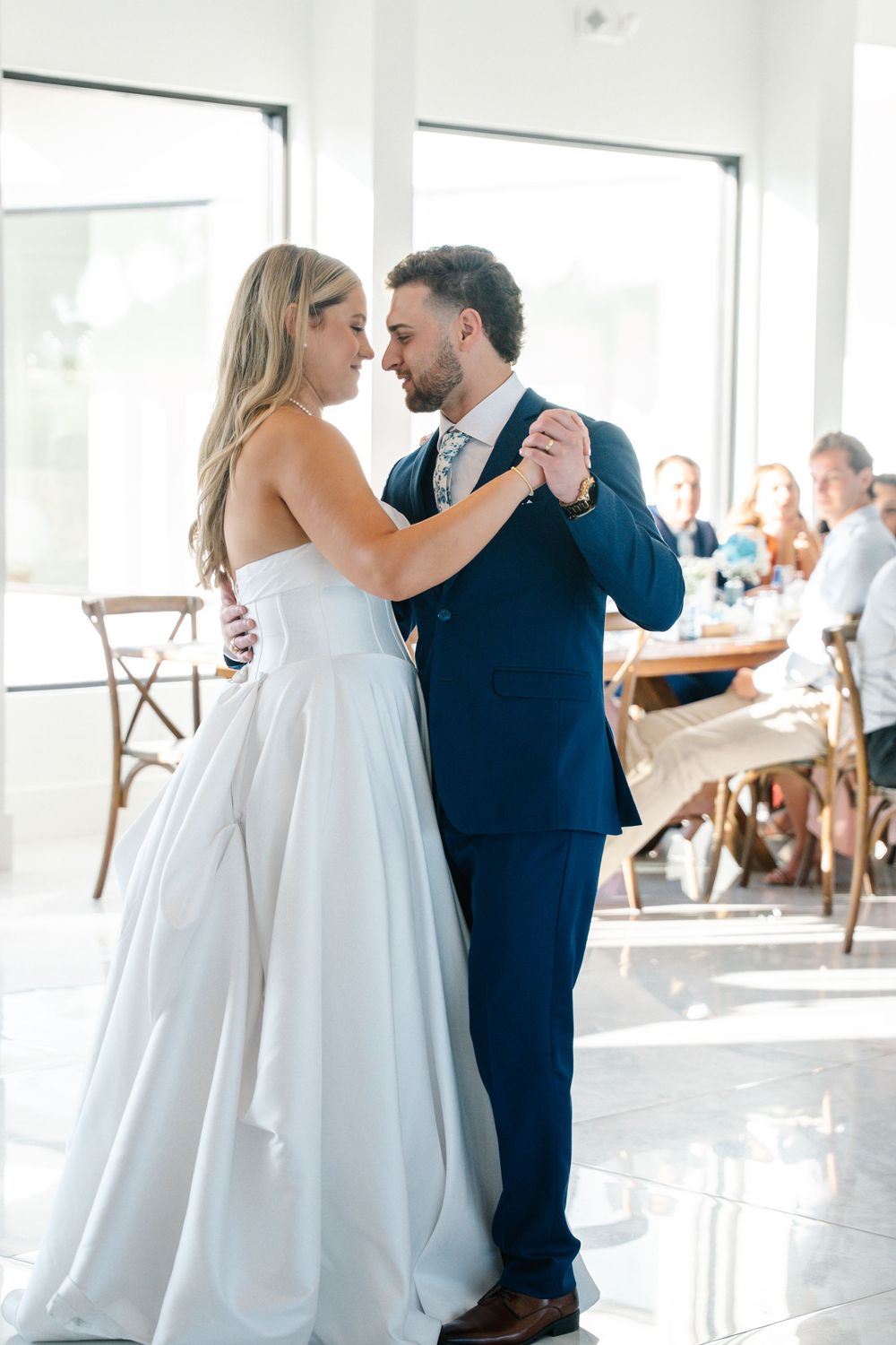 Bride and groom share their first dance in bright reception hall with guests watching.