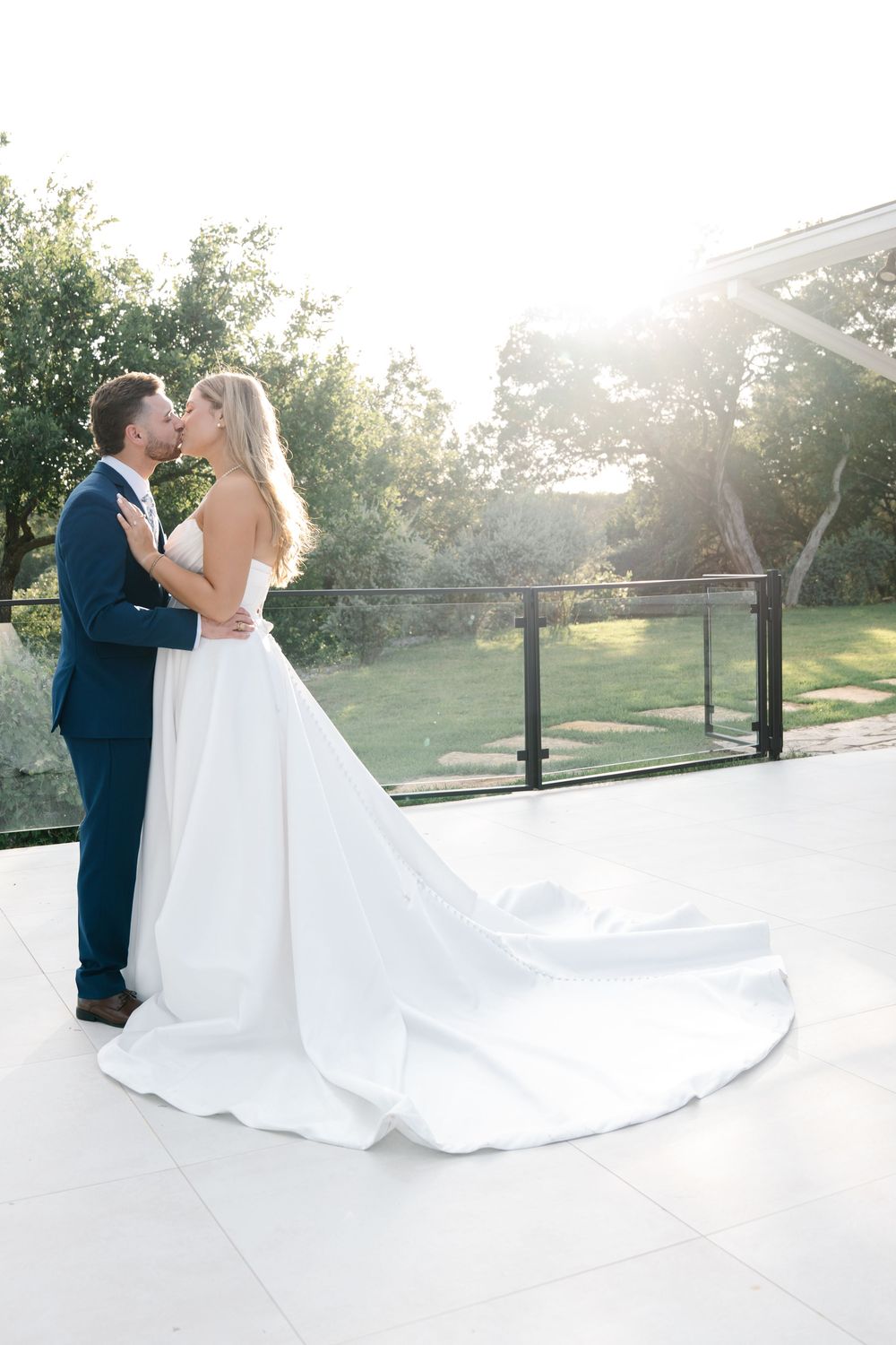 Bride and groom kissing on sunlit terrace with long cathedral train spread elegantly.