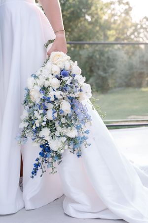 Bride holding cascading white and blue wedding bouquet with delphinium and roses against outdoor backdrop.
