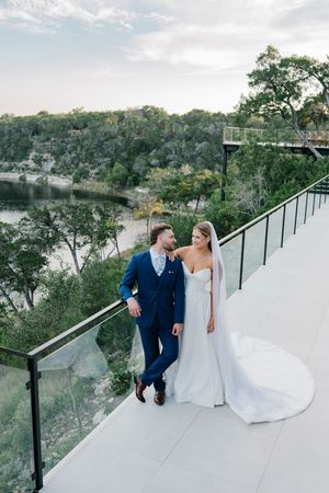 Bride and groom walking on terrace with panoramic river and forest views at dusk.