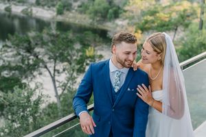 Smiling bride and groom in blue suit pose together on balcony with scenic river backdrop.