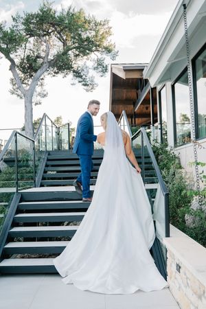 Bride in flowing white gown and groom in blue suit walk up outdoor stairs at wedding venue.