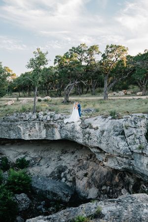 Bride and groom embrace on rocky cliff edge surrounded by trees at outdoor wedding.