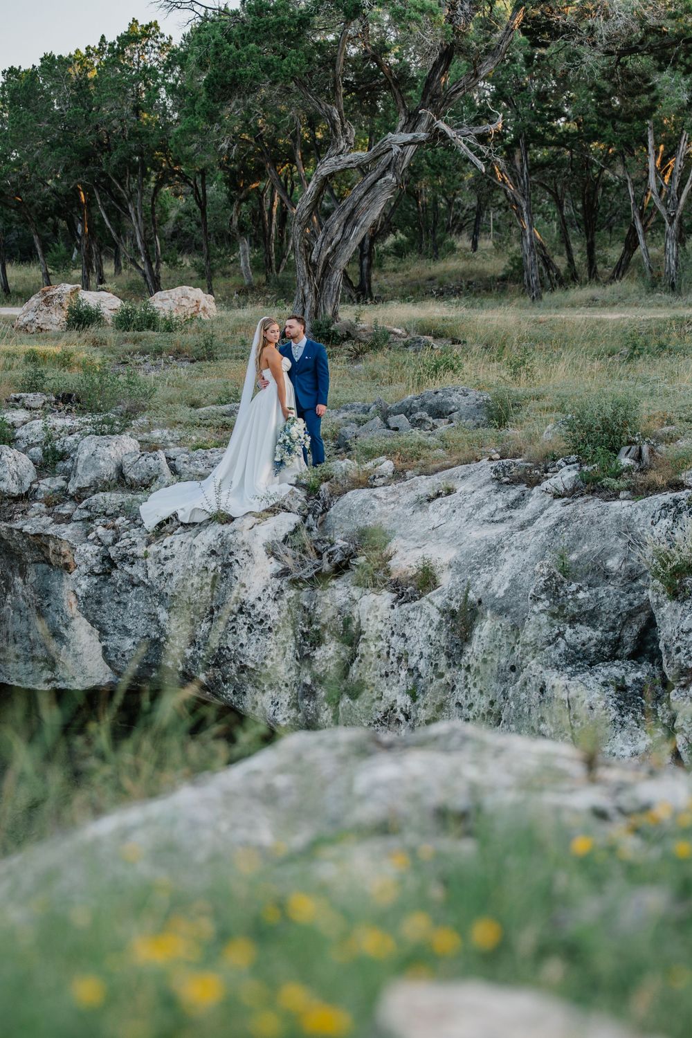 Bride in white gown with long veil and groom stand on rocky terrain in natural landscape.