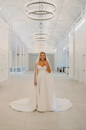 Elegant bride in strapless white ballgown with cathedral veil standing under crystal chandeliers in bright venue.