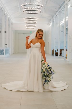 Smiling bride in strapless white ballgown holding blue and white cascading bouquet under crystal chandeliers.